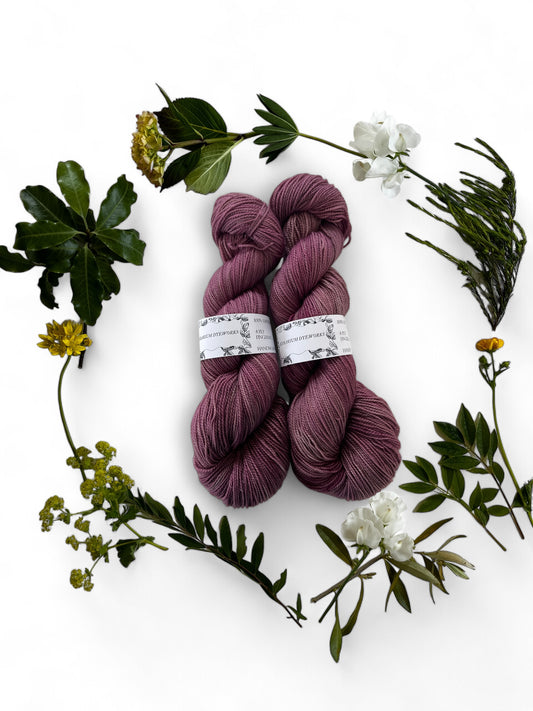 Two skeins of merino yarn in damson purple-pink dyed with Lac and Walnut and surrounded by a wreath of flowers and leaves